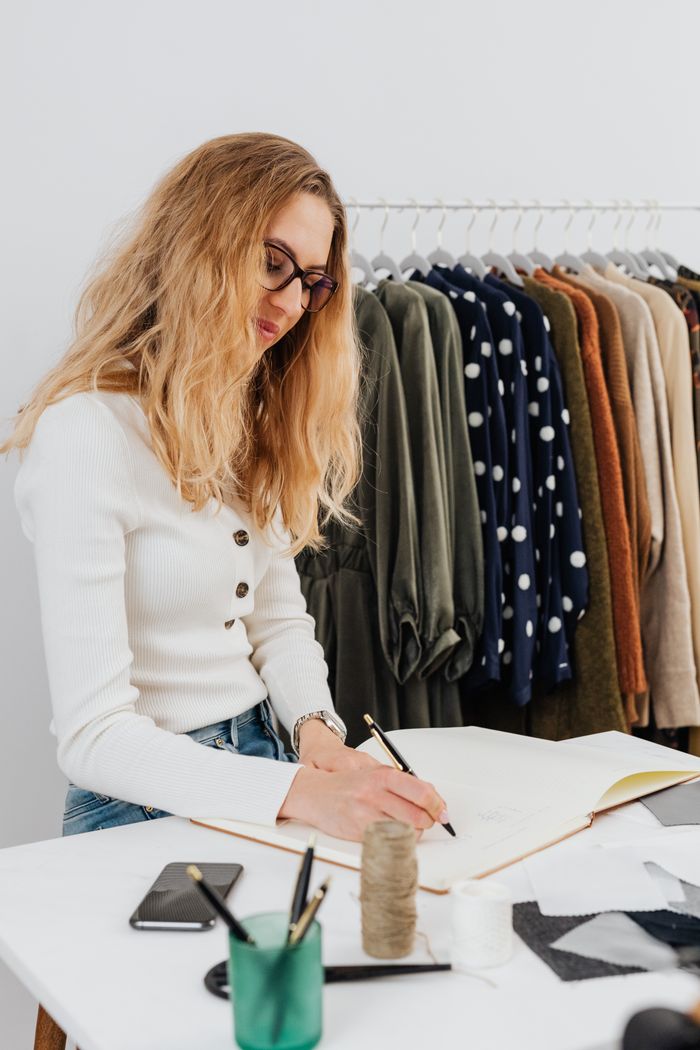 Woman creating fashion sketches in a clothing store with garments in the background.
