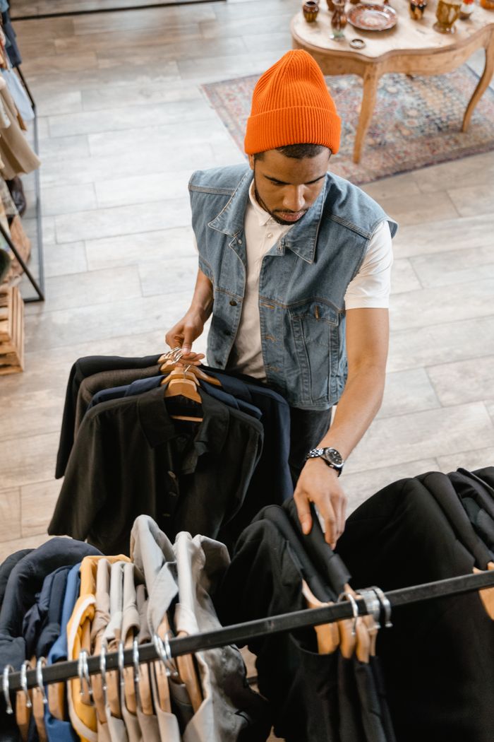 mobile-01 Man examines clothing in boutique wearing denim vest and orange beanie.